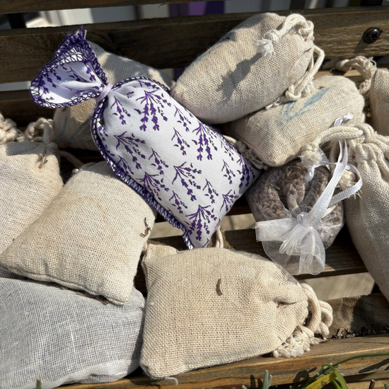 A pile of small burlap drawstring sachets and a lavender-patterned pillow resting on a wooden surface outdoors.